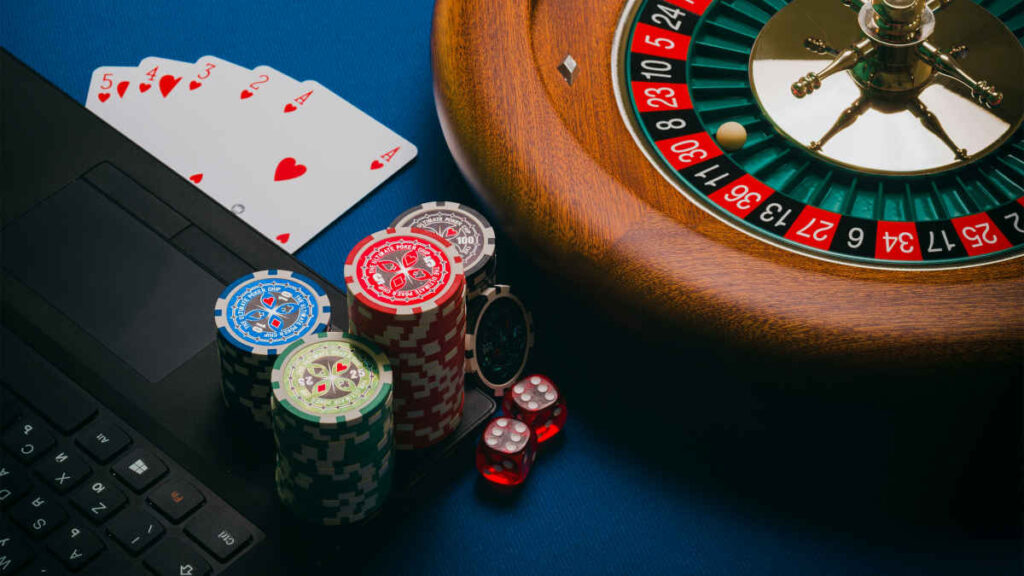 Casino table scene with poker chips, playing cards, roulette wheel, and a laptop keyboard on blue felt surface.